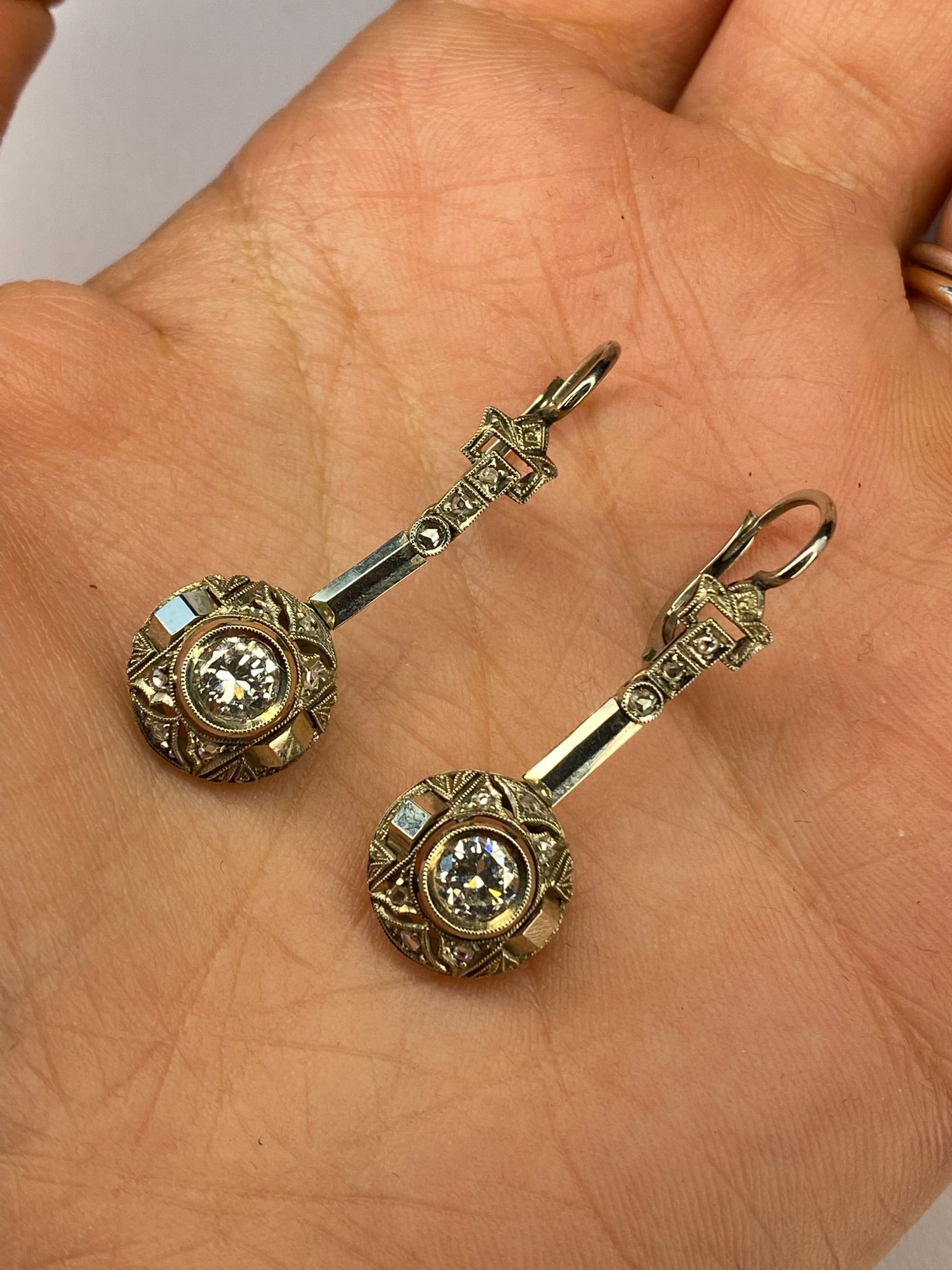 A pair of vintage-style gold-toned earrings with round bezel-set clear stones and ornate bar hooks, displayed on a person's palm.